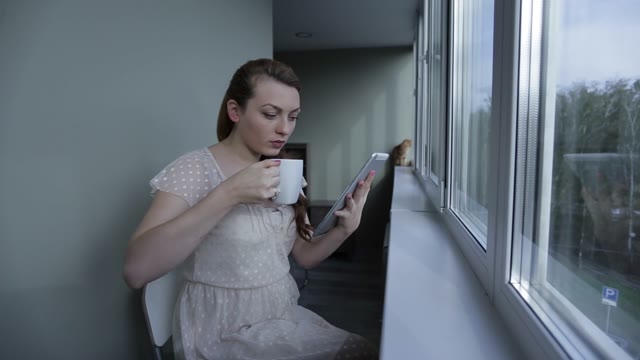 Girl With Tablet Sitting By The Window And Drink Of The Cup.