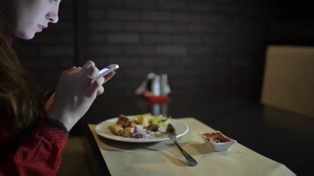 The Girl In The Red Dress Sits At A Table In The Restaurant And Use The Phone.
