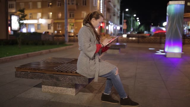 Young Woman Sitting On A Bench In The Street, And Enjoys The Tablet.