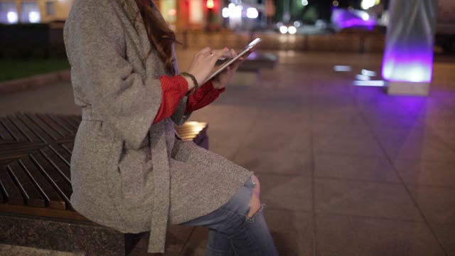 Young Girl In A Coat Sitting On A Bench In The Street, And Enjoys The Tablet.