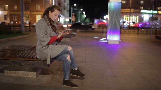 Young Girl Sitting On A Bench In The Street, And Enjoys The Tablet.