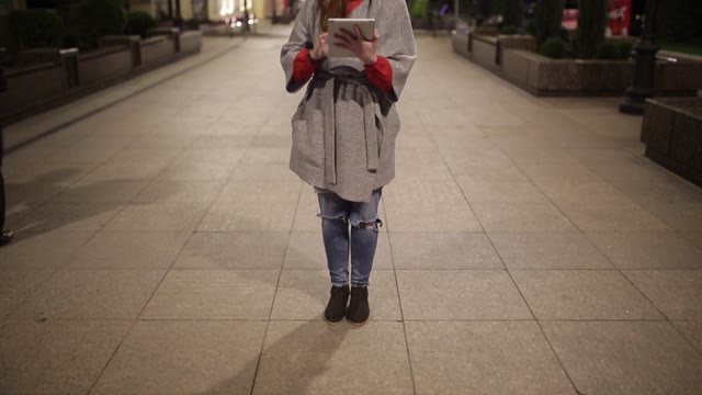 Young Girl In A Coat Is On The Boulevard With A Tablet In The Evening.