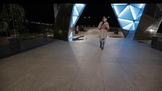 Cute Woman In Coat Walking Down The Street With A Tablet In The Evening.