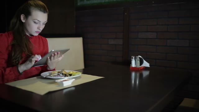 The Girl In The Red Dress Sits At A Table In The Restaurant And Use The Digital