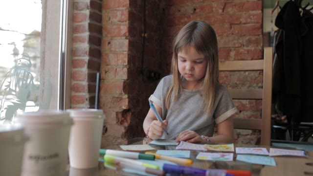 Smiling Little Girl Draws With Markers At A Table In A Coffee Shop