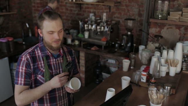 Young Guy With A Beard Behind The Counter In A Coffee Shop Writing On The Cup