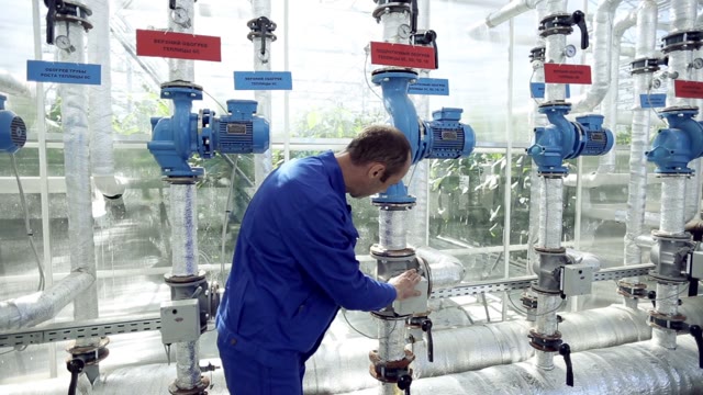 Engineer In Blue Overalls Repairing Pipework In The Greenhouse