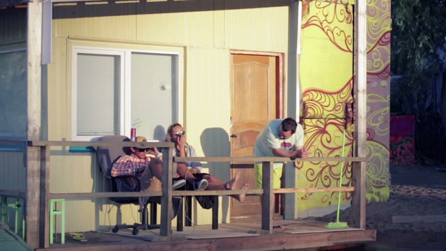 Three Young People Resting In The Sun Next To The House Near The Water