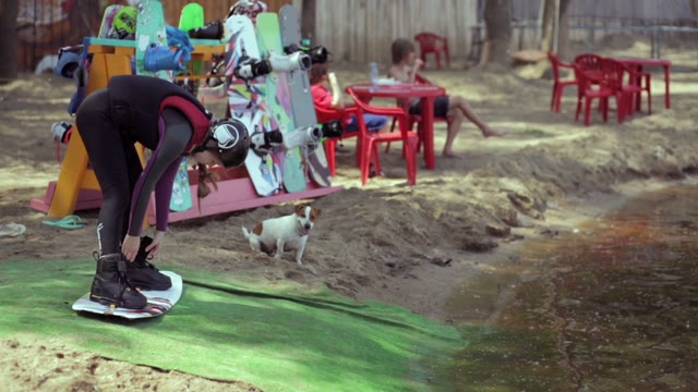 Beautiful Young Girl Fastens Buttons On Wakeboard. The Dog Runs Next.