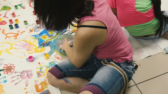 Girl Draws His Hands On His Face. Behind Pictures On The Floor.