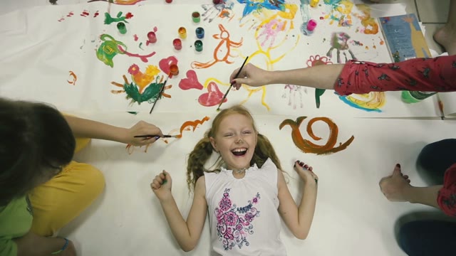 On The Floor Lies A Little Girl On Top Of Color Pictures, The Children Paint.