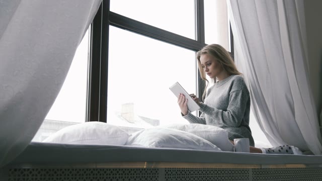Young Girl Sitting On A Window Sill And Enjoys The Tablet. Drinking From A Glass