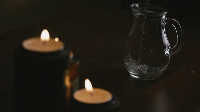 Pour Water In A Pitcher In The Foreground Lit Candles. Close-up. Sow Motion