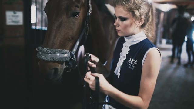 Beautiful Blond Girl Horsewoman Puts On The Reins Of Her Horse In The Stable