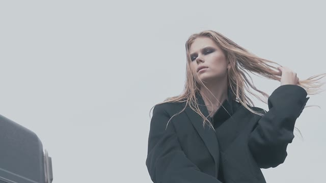 Girl With Long Hair In Black Coat Poses For A Photo On A Background Cloudy Sky
