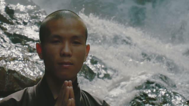 Monk Stands Near The Waterfall Close-up