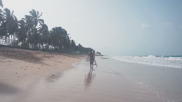 A Nice And Attractive Dark-haired Surfer Walks Along The Tropical Beach