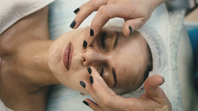 Employee Of The Spa Salon Applies A Thin Layer Of Cream Mask On The Face Of A